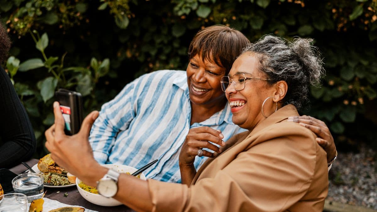 Happy woman taking selfie with senior woman through smart phone sitting at dining table in back yard.