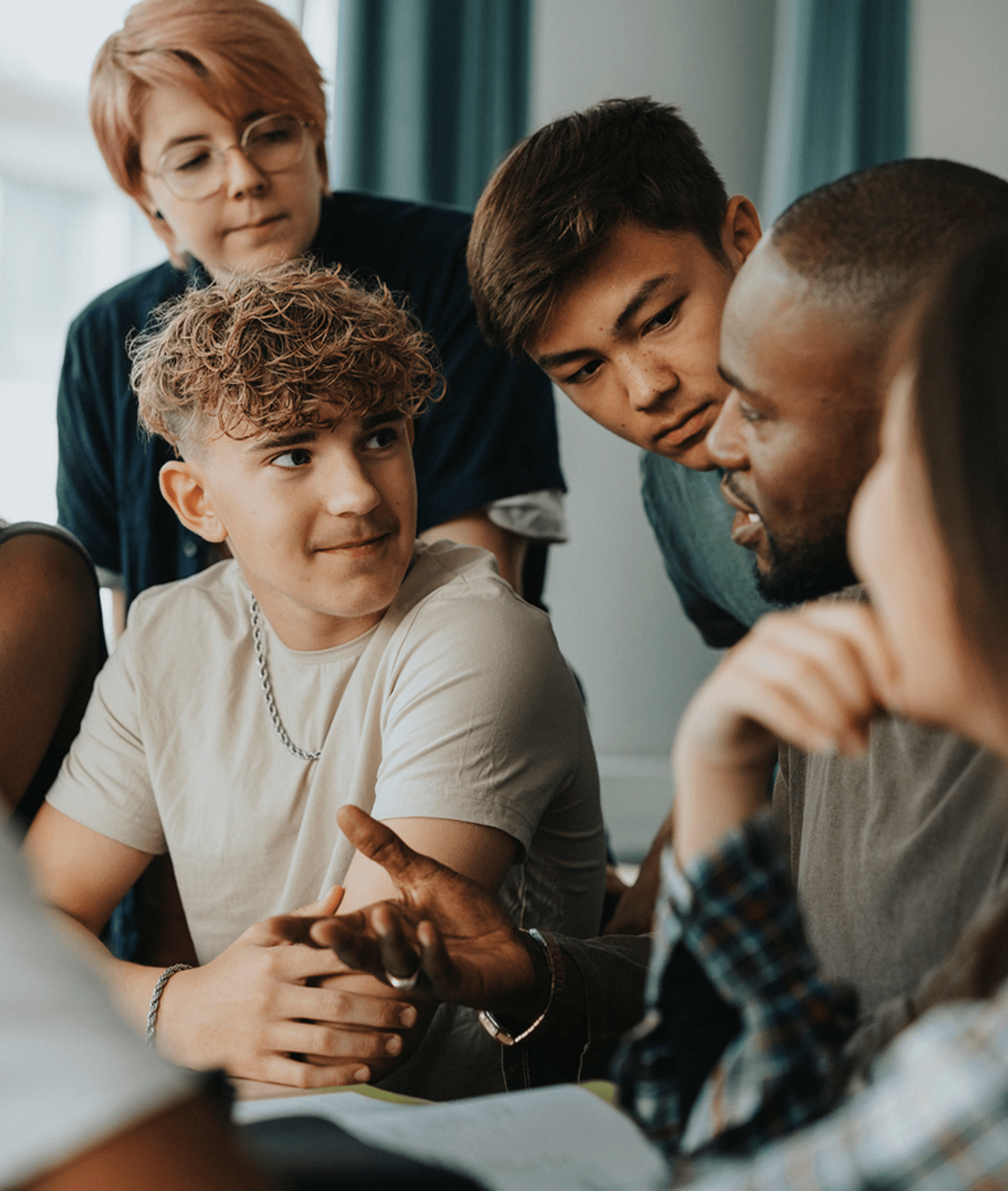 Teenage boys and girls with non-binary person in discussion with a male teacher around a table in a classroom.