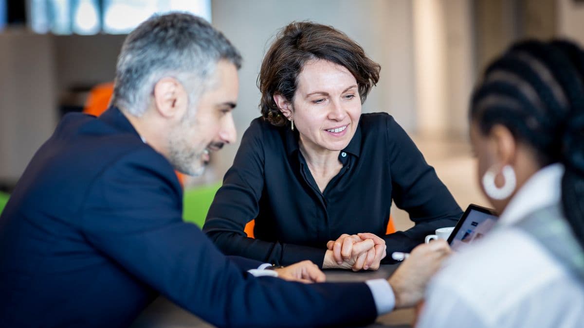 A man and two women sat at a table around an iPad in discussion.